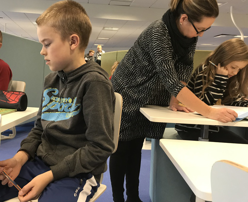 A teacher helps a fifth-grade student in a classroom and a boy sits at his desk.