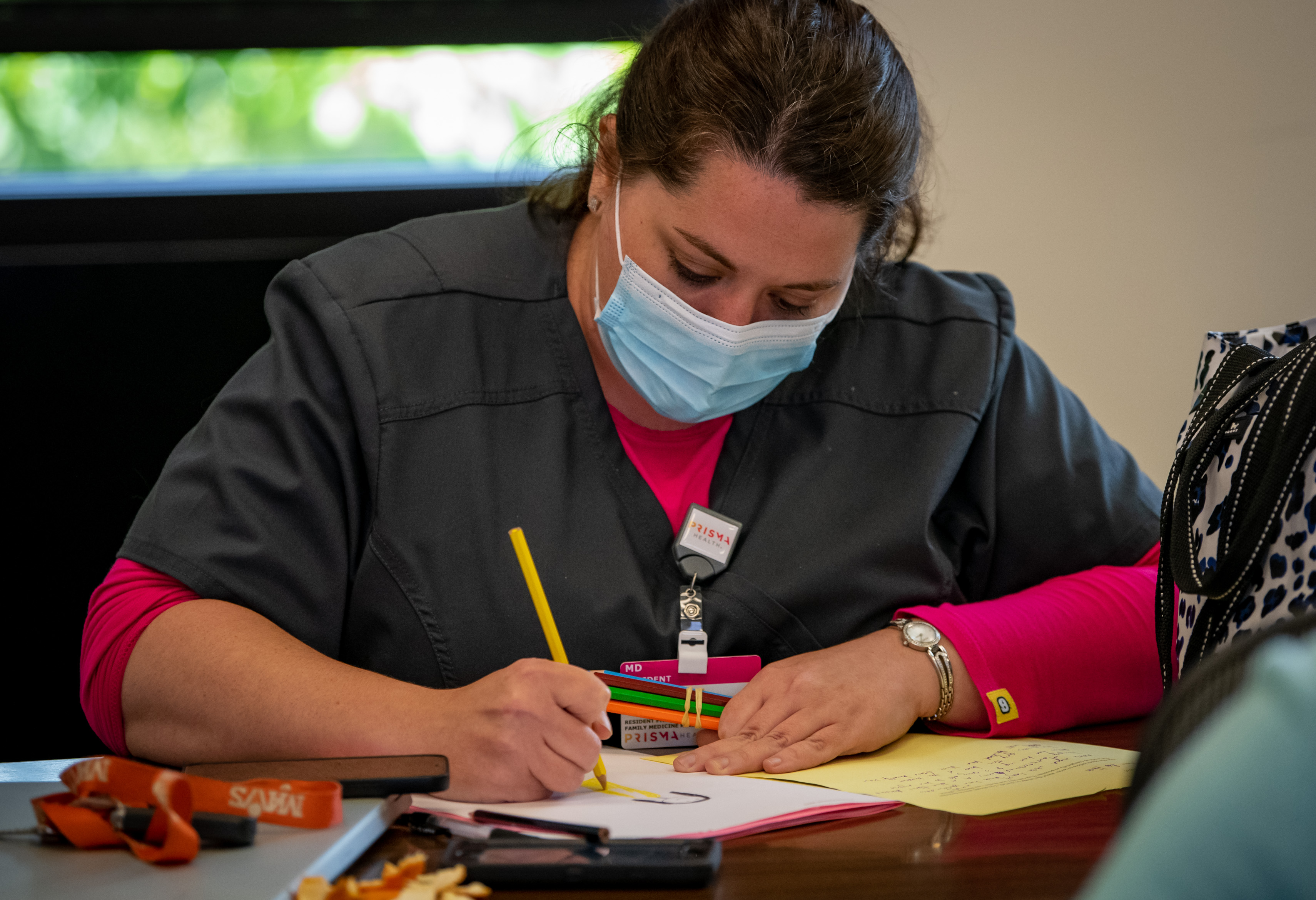 A woman in scrubs and a mask draws with colored pencils.